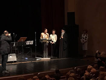 On the stage on the podium is a girl making a speech, a woman and a priest standing next to her, the photographer taking pictures of her and the audience sitting below watching her.